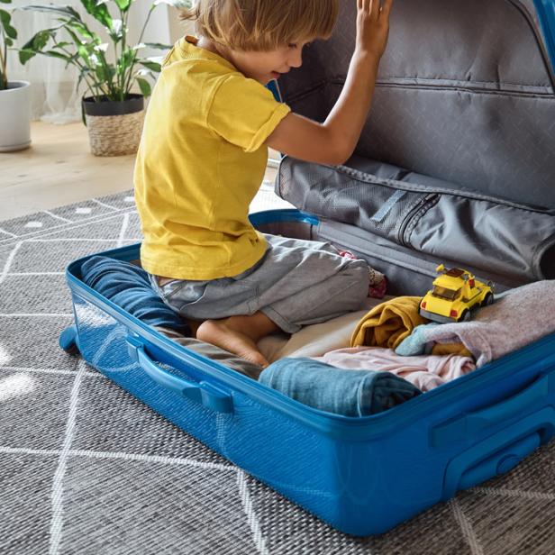 Ein Junge sitzt in einem blauen Koffer, umgeben von Kleidung und einem Spielzeugauto.