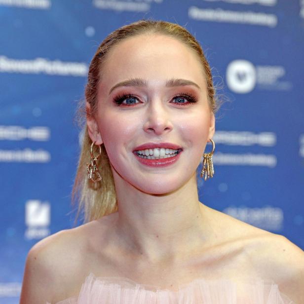 Porträt einer lächelnden Frau mit blonden Haaren und Ohrringen vor blauem Hintergrund.
