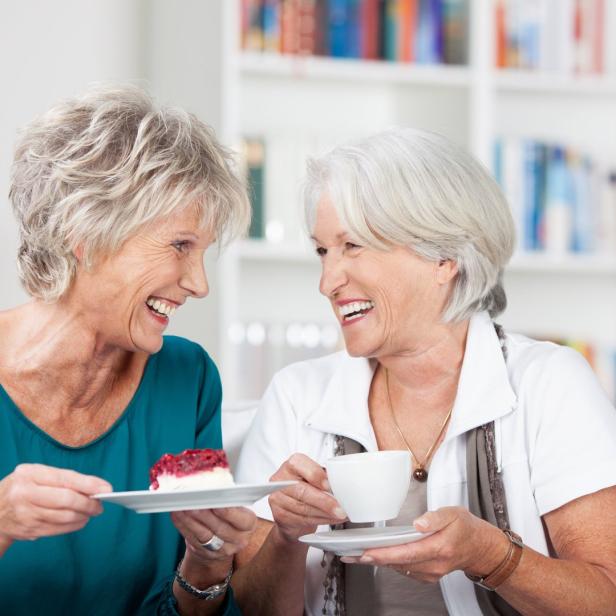 Zwei ältere Frauen lachen und halten jeweils ein Stück Kuchen und eine Tasse Kaffee.