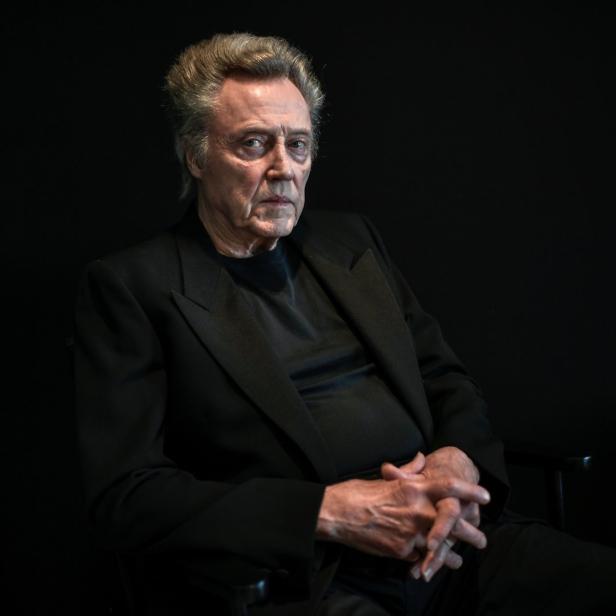 Christopher Walken sitzt in einem schwarzen Anzug vor einem dunklen Hintergrund.