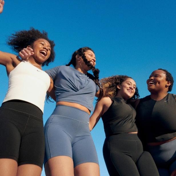 Vier Frauen in Sportkleidung von Nike stehen zusammen und lachen vor blauem Himmel.
