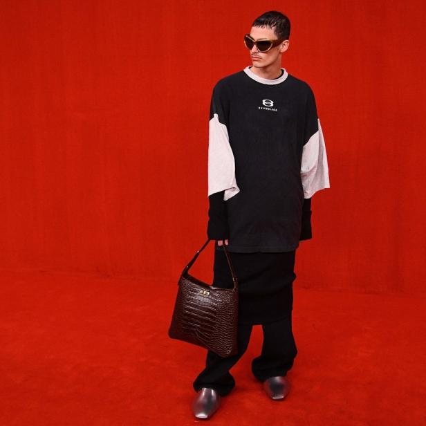 Ein Mann mit Sonnenbrille, Oversize-Shirt und Handtasche vor rotem Hintergrund.