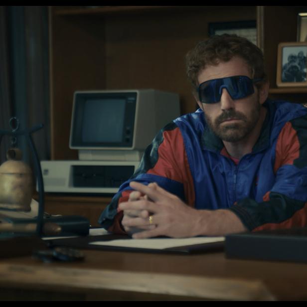 Ben Affleck sitzt mit Sonnenbrille an einem Schreibtisch mit einem alten Computer.