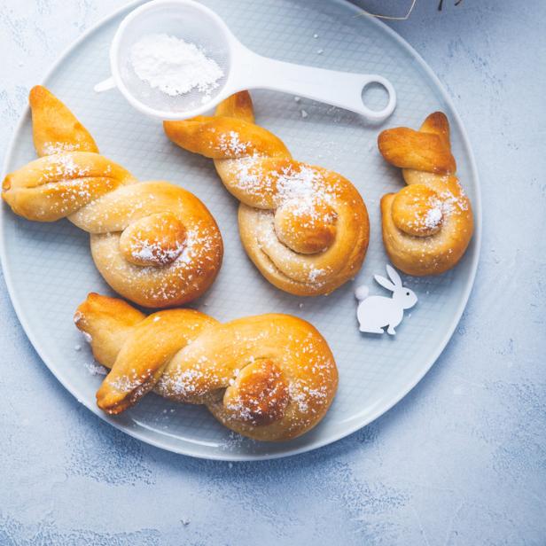 Ein Teller mit süßen, hasenförmigen Brötchen, bestäubt mit Puderzucker, neben einem Nest mit bunten Ostereiern.