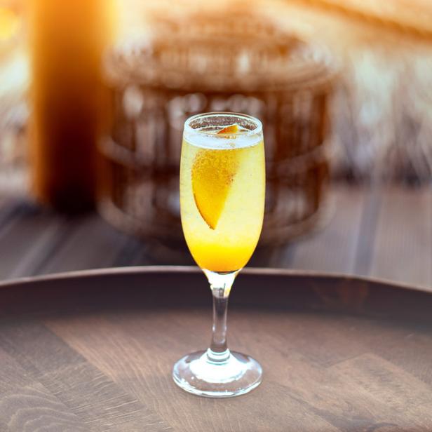 Ein Glas Sekt mit einem Pfirsichstück auf einem Holztablett.