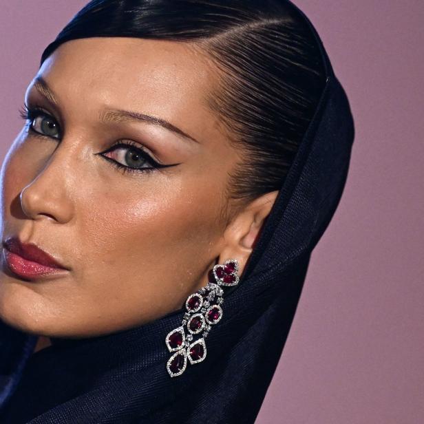 Nahaufnahme von Bella Hadid mit einem schwarzen Kopftuch und einem roten Ohrring vor einem rosa Hintergrund.