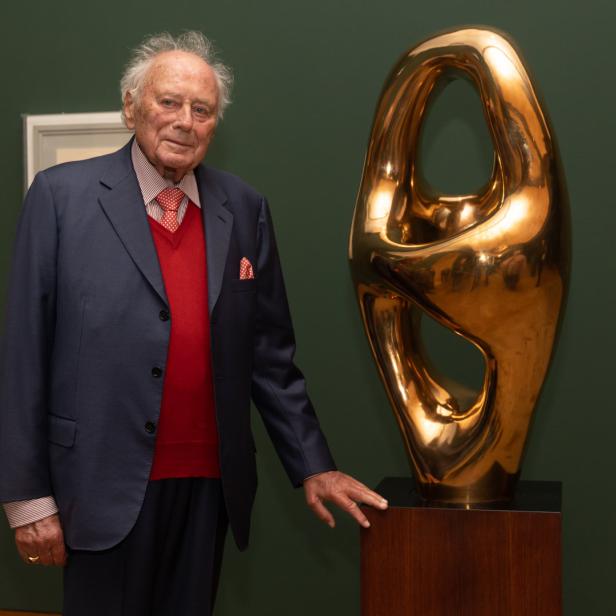 Ein älterer Mann posiert neben einer abstrakten, goldenen Skulptur.