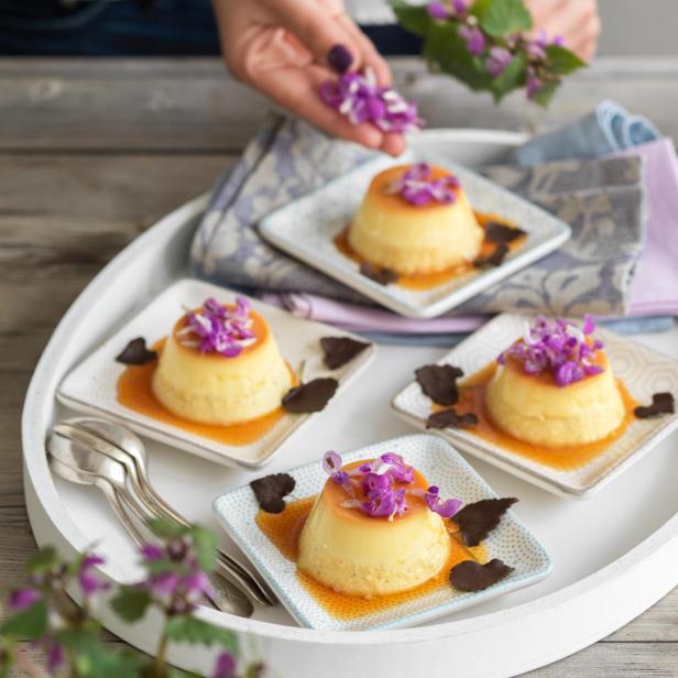 Vier Puddingförmchen mit Karamell und lila Blüten auf einem Tablett angerichtet.
