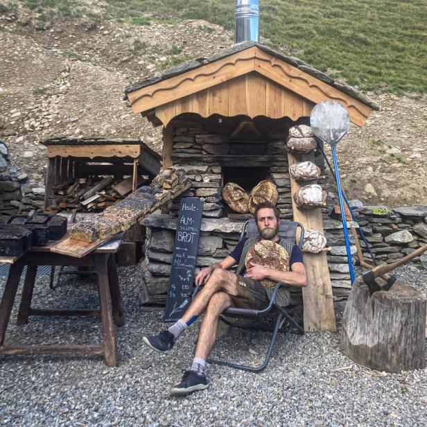 Ein Mann in Lederhosen sitzt vor einem Steinofen und hält ein Brotlaib.