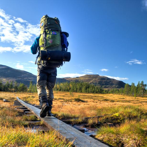 Ein Wanderer mit großem Rucksack überquert einen Holzsteg in einer Moorlandschaft.