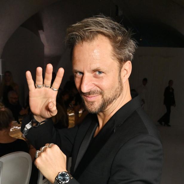 Ein Mann mit Anzug und mehreren Ringen zeigt mit der Hand eine Vier.