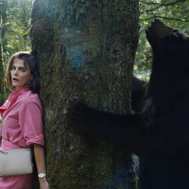 Eine Frau in einem Wald lehnt ängstlich an einem Baum, während sich ein Bär nähert.