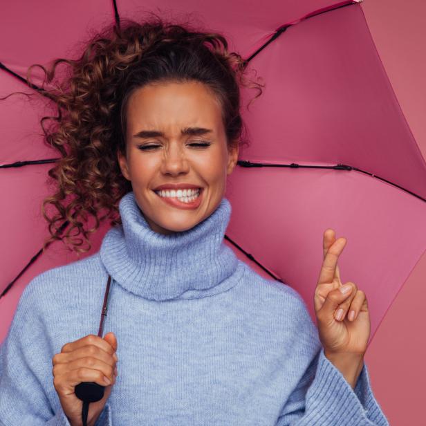 Eine Frau mit gekreuzten Fingern hält einen rosa Regenschirm vor einem rosa Hintergrund.