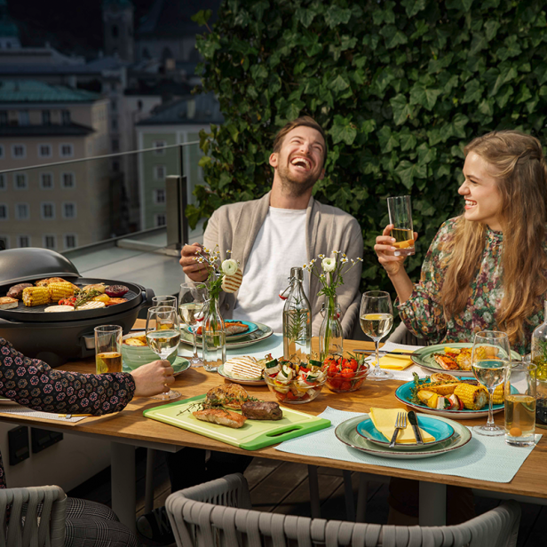 Drei Freunde sitzen auf einer Dachterrasse beim Grillen und genießen die Aussicht.