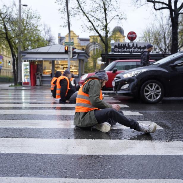 Mehrere Demonstranten in Warnwesten sitzen auf einem nassen Zebrastreifen und blockieren den Verkehr.