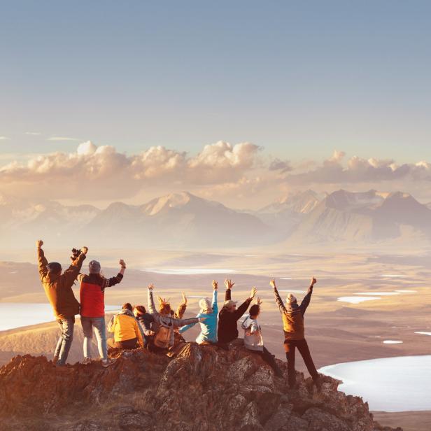 Eine Gruppe von Menschen jubelt auf einem Felsen mit Blick auf eine weite Landschaft mit Bergen und Seen.