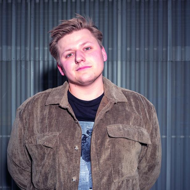 Ein junger Mann mit blonden Haaren und brauner Cordjacke vor einer grauen Wand.