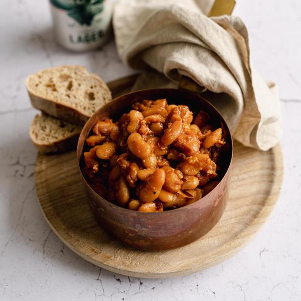 Ein Topf mit Baked Beans steht auf einem Holzteller, dazu Brot und ein Bier.
