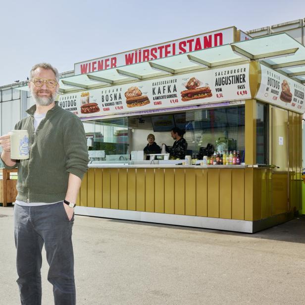 Ein Mann mit Bierkrug steht vor einem „Wiener Würstelstand“.