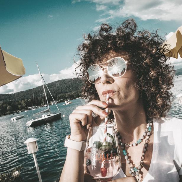 Eine Frau mit lockigem Haar trinkt mit Strohhalm ein Getränk an einem See.