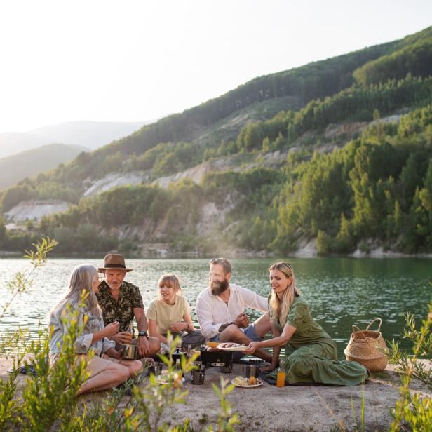 Eine Familie grillt am Ufer eines Sees vor einer bewaldeten Hügellandschaft.