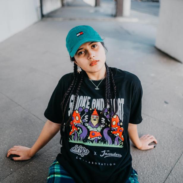 Eine junge Frau mit Zöpfen und einem T-Shirt mit Pilzmotiv sitzt auf dem Boden.