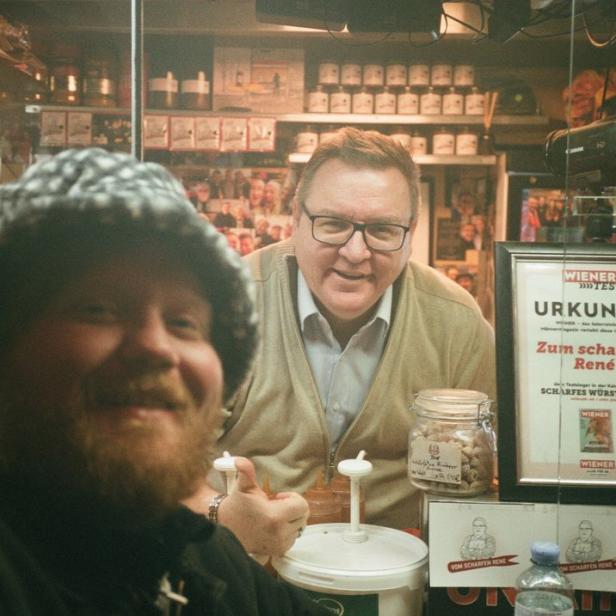Ein Mann mit Bart und Mütze vor einem Hotdog-Stand mit einem Porträt von René im Hintergrund.