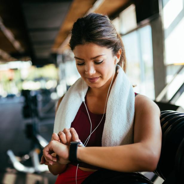 Eine Frau mit einem Handtuch um den Hals schaut auf ihre Smartwatch im Fitnessstudio.