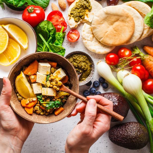 Eine Person hält eine Schüssel mit Tofu, Gemüse und Linsen in der Hand. Um die Schüssel herum sind weitere Zutaten angerichtet.