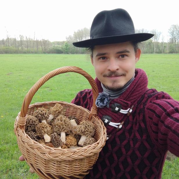Ein Mann mit Hut präsentiert einen Korb voller Morcheln auf einer Wiese.