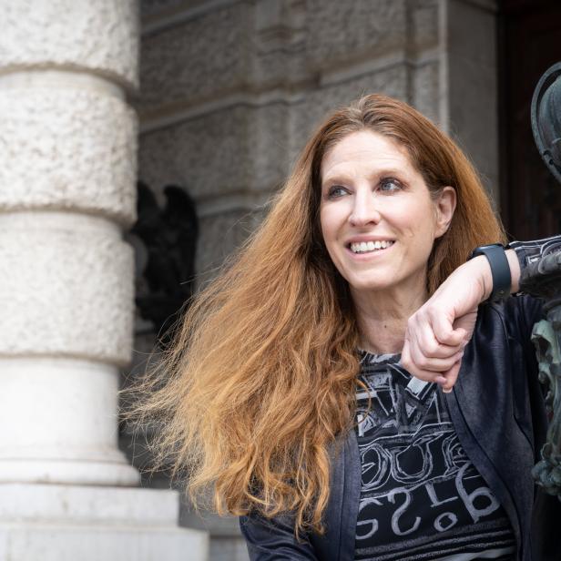 Eine Frau mit langen, roten Haaren lehnt an einer verzierten Laterne vor einem Gebäude.