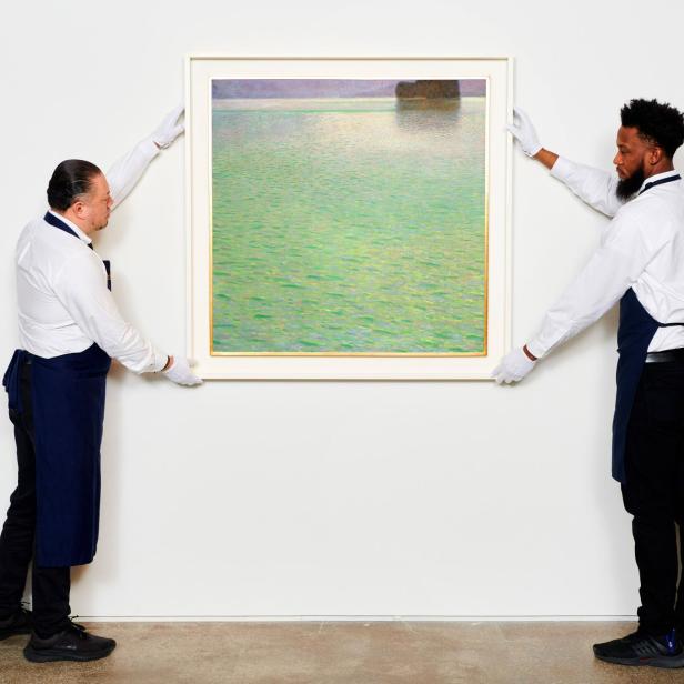 Zwei Personen in Arbeitskleidung hängen ein Landschaftsgemälde an eine weiße Wand.