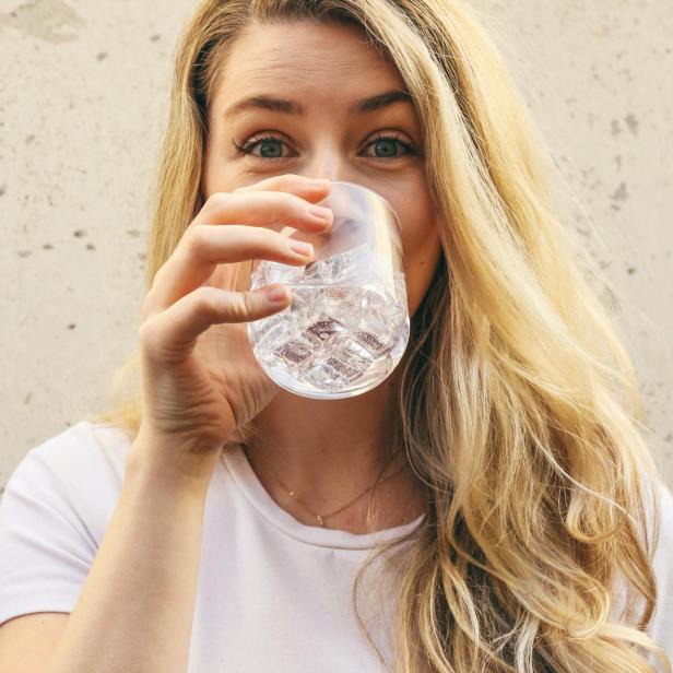 Eine Frau mit blonden Haaren trinkt ein Glas Wasser mit Eiswürfeln.