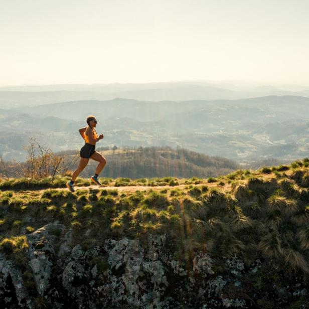 Eine Frau läuft auf einem Bergpfad mit Blick auf eine hügelige Landschaft.