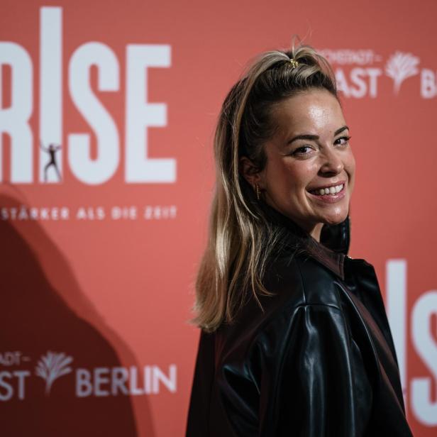 Eine lächelnde Frau mit Pferdeschwanz steht vor rotem ARISE-Hintergrund mit PALAST BERLIN-Schriftzug.