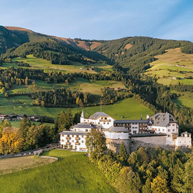 Ein Schloss thront auf einem Hügel, umgeben von grünen Wiesen und bewaldeten Bergen.