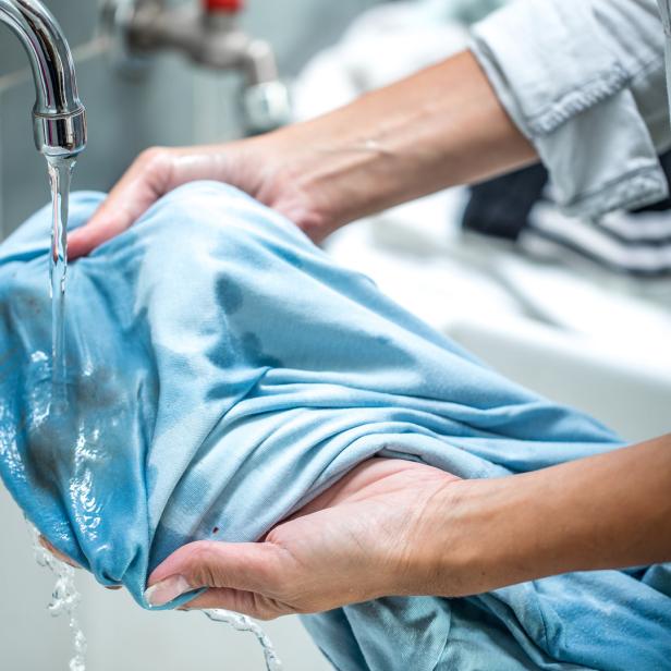Eine Person wäscht ein blaues Hemd mit Flecken unter einem Wasserhahn.