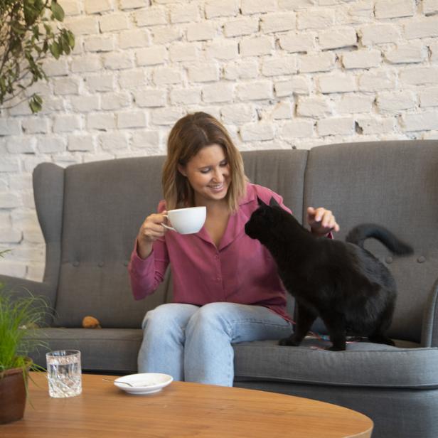 Eine Frau sitzt auf einem Sofa, hält eine Tasse und streichelt eine schwarze Katze.