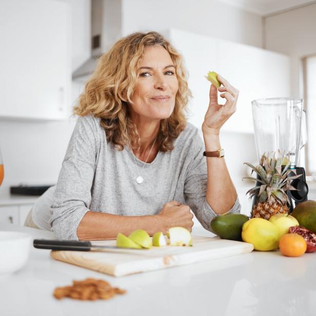 Eine Frau in einer Küche isst ein Stück Apfel vor einem Mixer und frischem Obst.