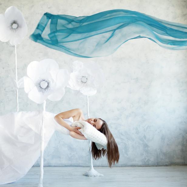 Eine Frau in einem weißen Kleid liegt auf einer Schaukel, umgeben von Papierblumen und einem blauen Tuch.