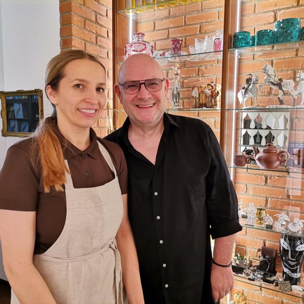 Eine Frau mit Schürze und ein Mann stehen vor einem Regal mit Glaswaren.