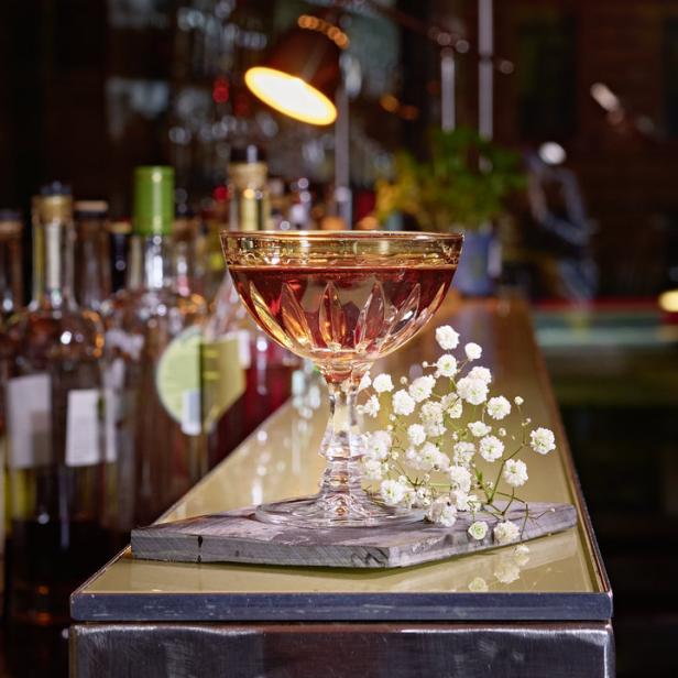 Ein Cocktail in einem Kristallglas mit Goldrand steht auf einer Bar, dekoriert mit Schleierkraut.