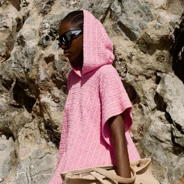 Eine Frau mit Sonnenbrille und einem rosa Givenchy-Strandponcho mit passender Tasche vor einem Felsen.