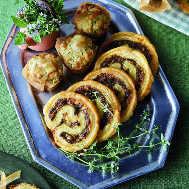 Ein silbernes Tablett mit Muffins, Blätterteigschnecken und einem kleinen Topf mit Kräutern.