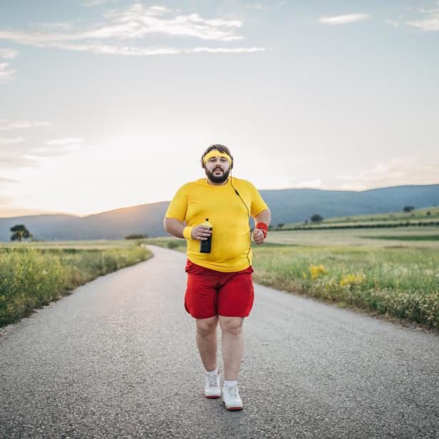 Ein Mann mit gelbem Stirnband joggt mit Kopfhörern und einer Wasserflasche auf einer Landstraße.