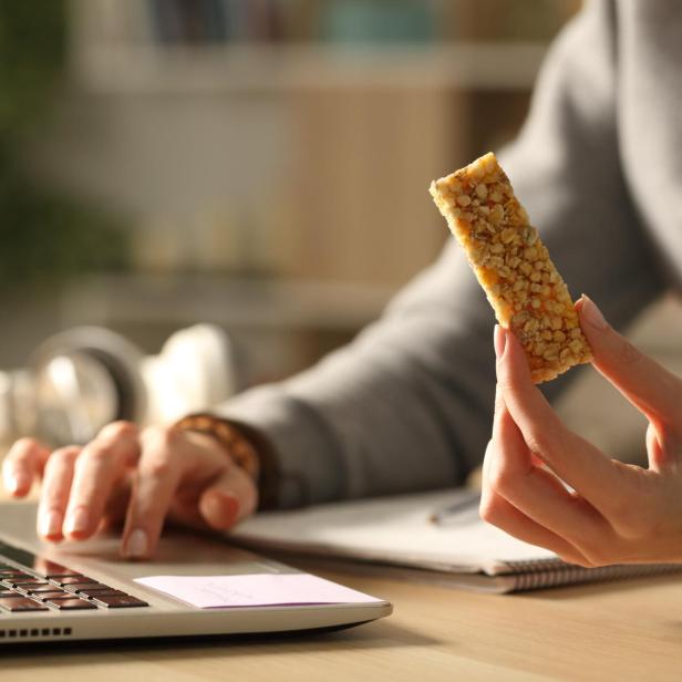 Eine Person arbeitet am Laptop und hält einen Müsliriegel in der Hand.
