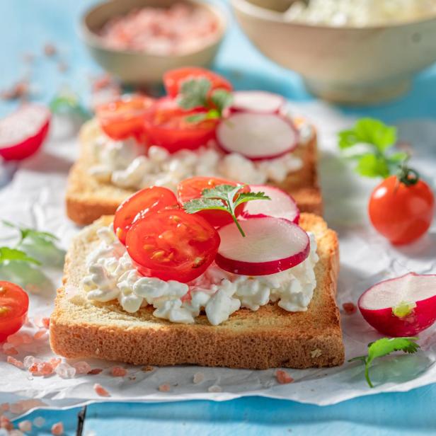 Zwei Toasts mit Hüttenkäse, Tomaten und Radieschen auf blauem Untergrund.