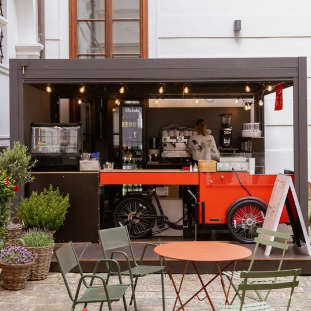 Ein mobiler Kaffeestand mit einer Frau im Hintergrund, umgeben von Tischen und Pflanzen.