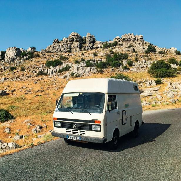 Ein weißer VW-Camper fährt auf einer Straße durch eine felsige Landschaft.