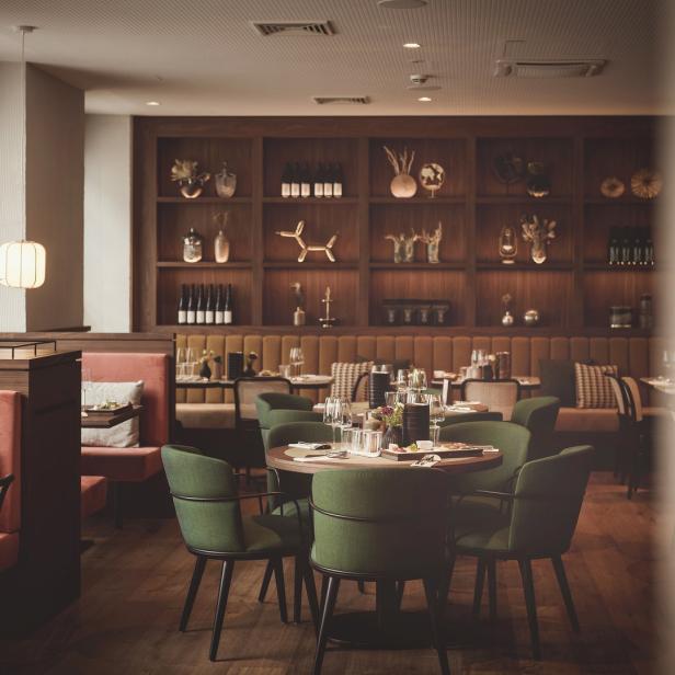 Ein elegantes Restaurant mit Tischen, grünen Stühlen und einer Wand mit dekorativen Objekten.